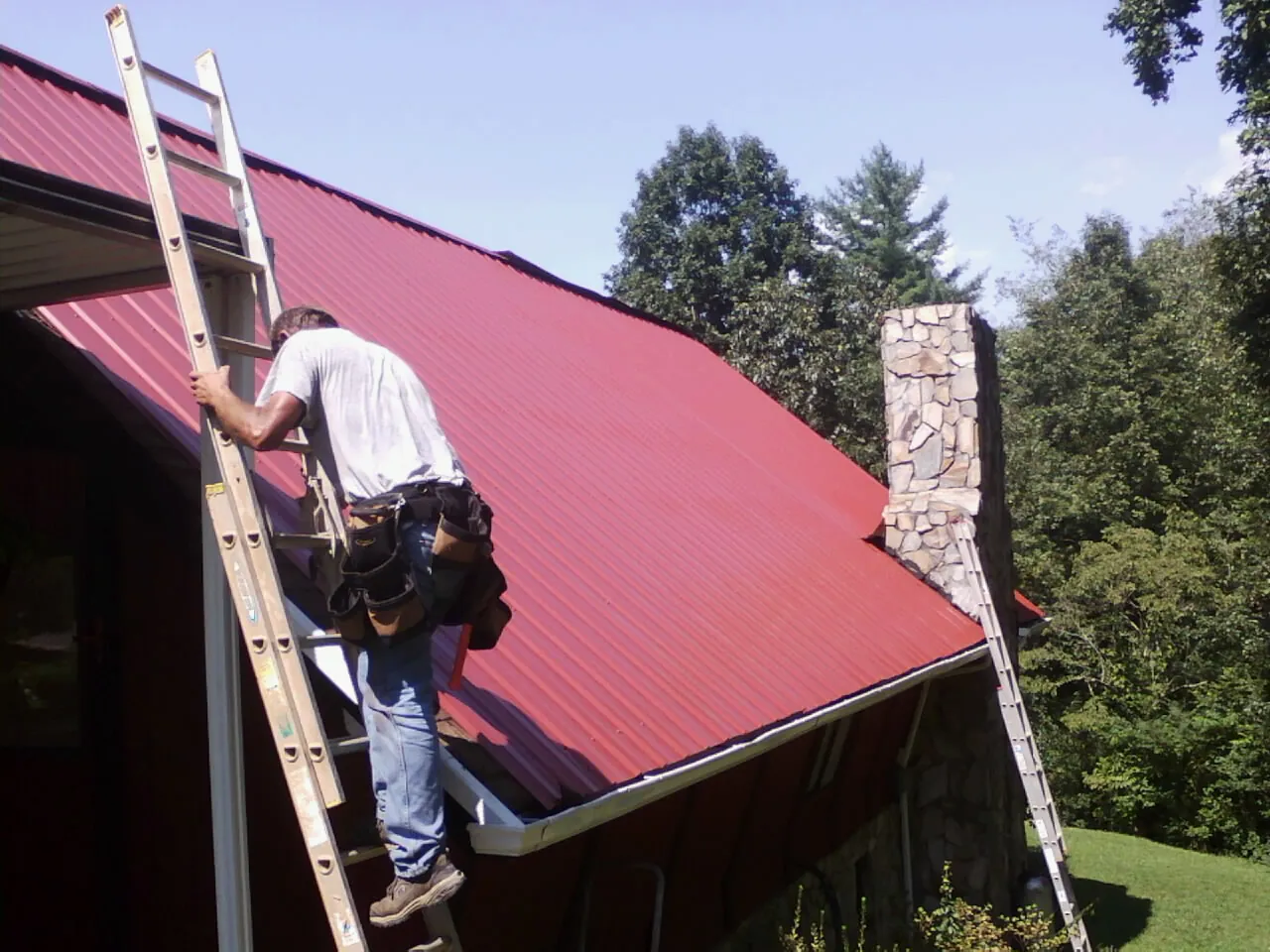 Roofer on a ladder beside a red metal roof and stone chimney — Soffit & Fascia Repair by Roofing Brothers in Walker
